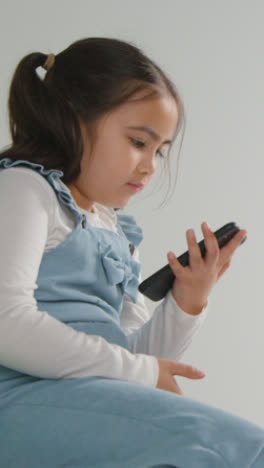 Vertical-Video-Studio-Shot-Of-Young-Girl-On-ASD-Spectrum-Gaming-On-Mobile-Phone-Against-White-Background